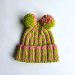 Knitted hat with pink and green stripes and two pom-poms on a light gray background -- pattern by Isknit, yarn by Global Backyard (Polly + Blushing Peach).