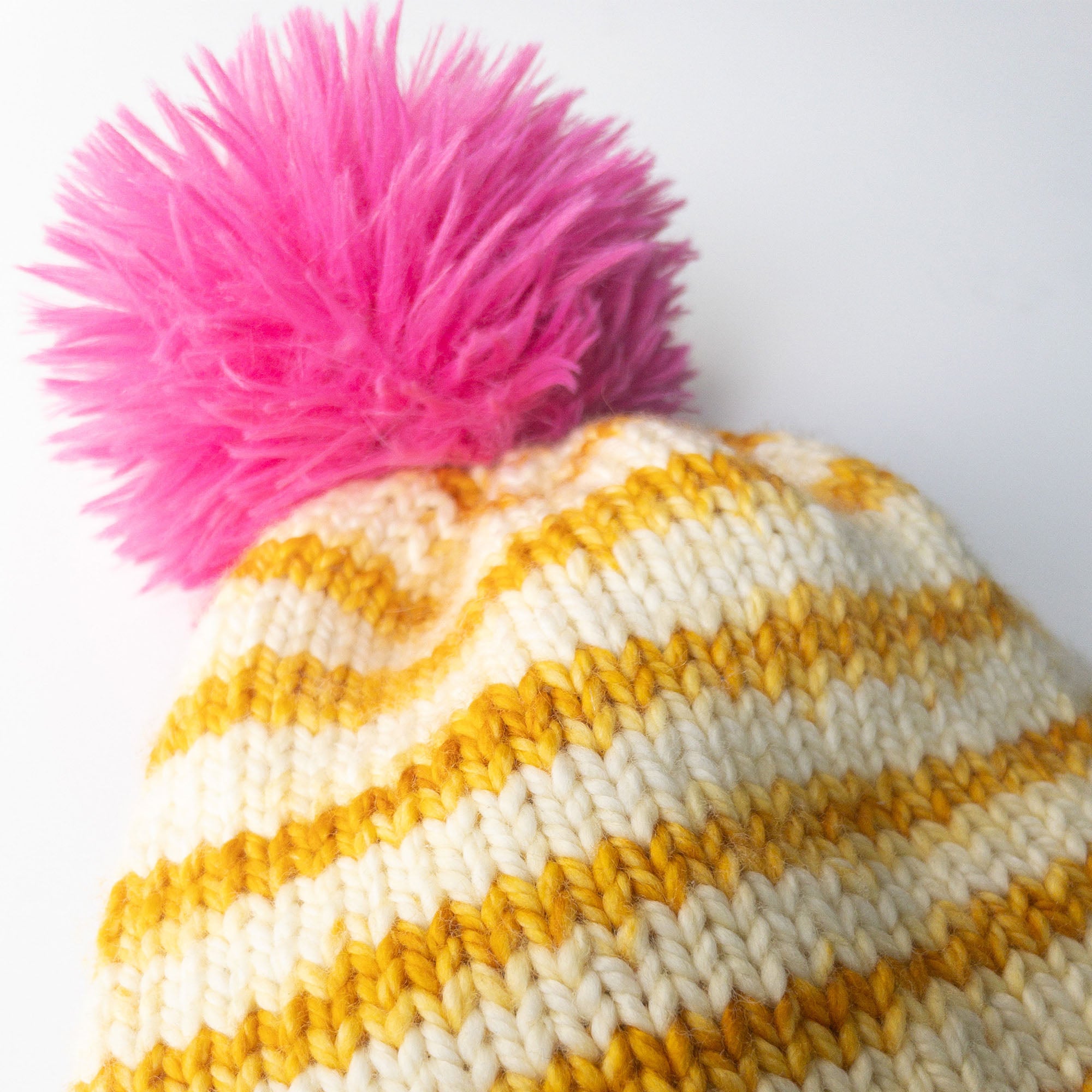 one-skein knitting project hat kit in vanilla cream and butterscotch (mustard) yellow that self-stripes on the free knitting hat pattern we recommend; also has a fuschia pink faux fur pompom topping it off