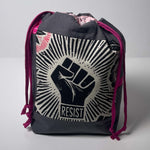 A dark gray drawstring bag featuring a large black and off-white raised fist graphic with the word 'RESIST' on the front, pink drawstrings, and a pink patterned interior lining, photographed against a neutral gray background.