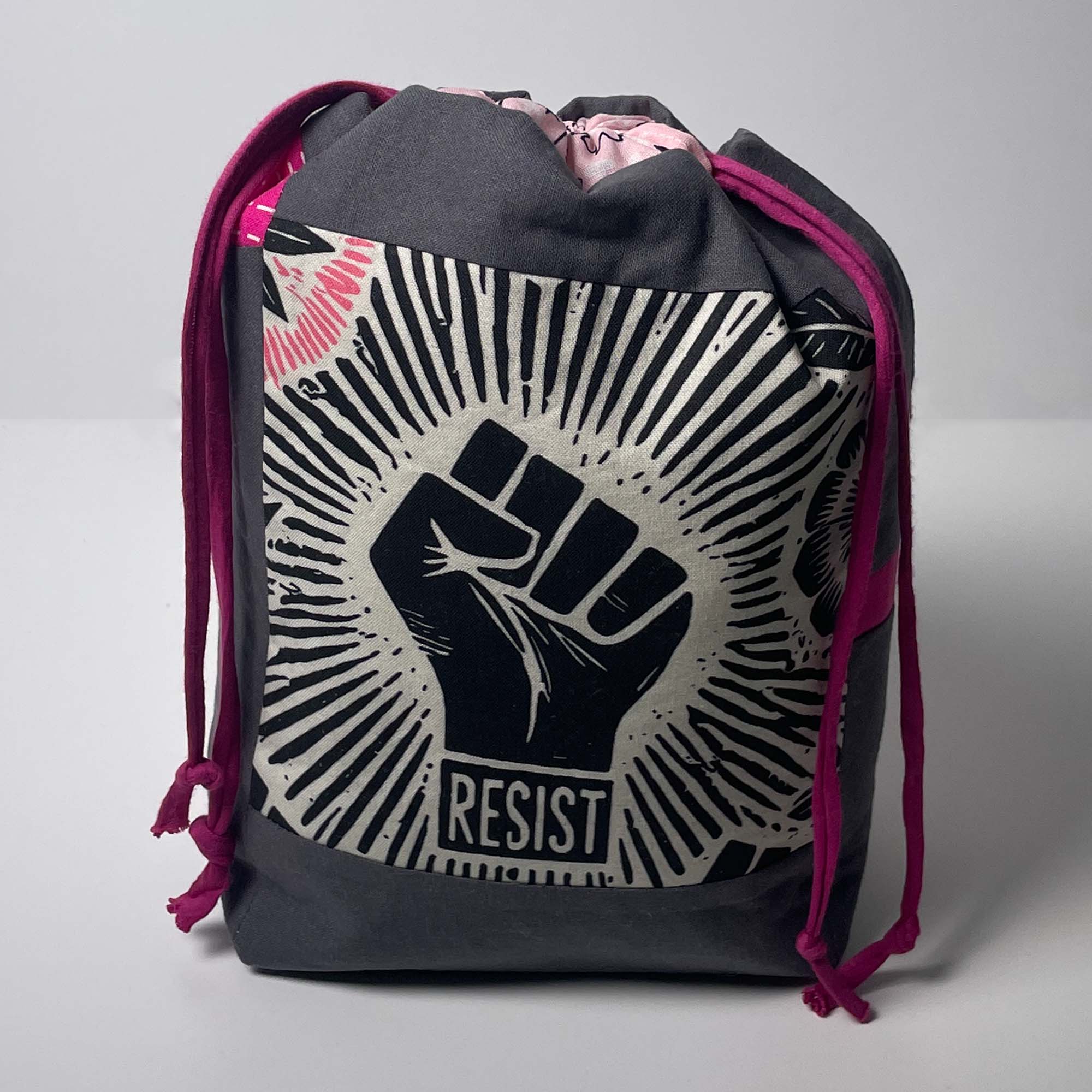 A dark gray drawstring bag featuring a large black and off-white raised fist graphic with the word 'RESIST' on the front, pink drawstrings, and a pink patterned interior lining, photographed against a neutral gray background.