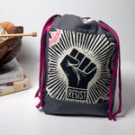 A dark gray drawstring backpack with a prominent black and white graphic of a raised fist and the word 'RESIST' on the front, accented with magenta drawstring cords, set against a light gray background with a decorative book and wooden bowl on the left.