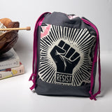 A dark gray drawstring backpack with a prominent black and white graphic of a raised fist and the word 'RESIST' on the front, accented with magenta drawstring cords, set against a light gray background with a decorative book and wooden bowl on the left.