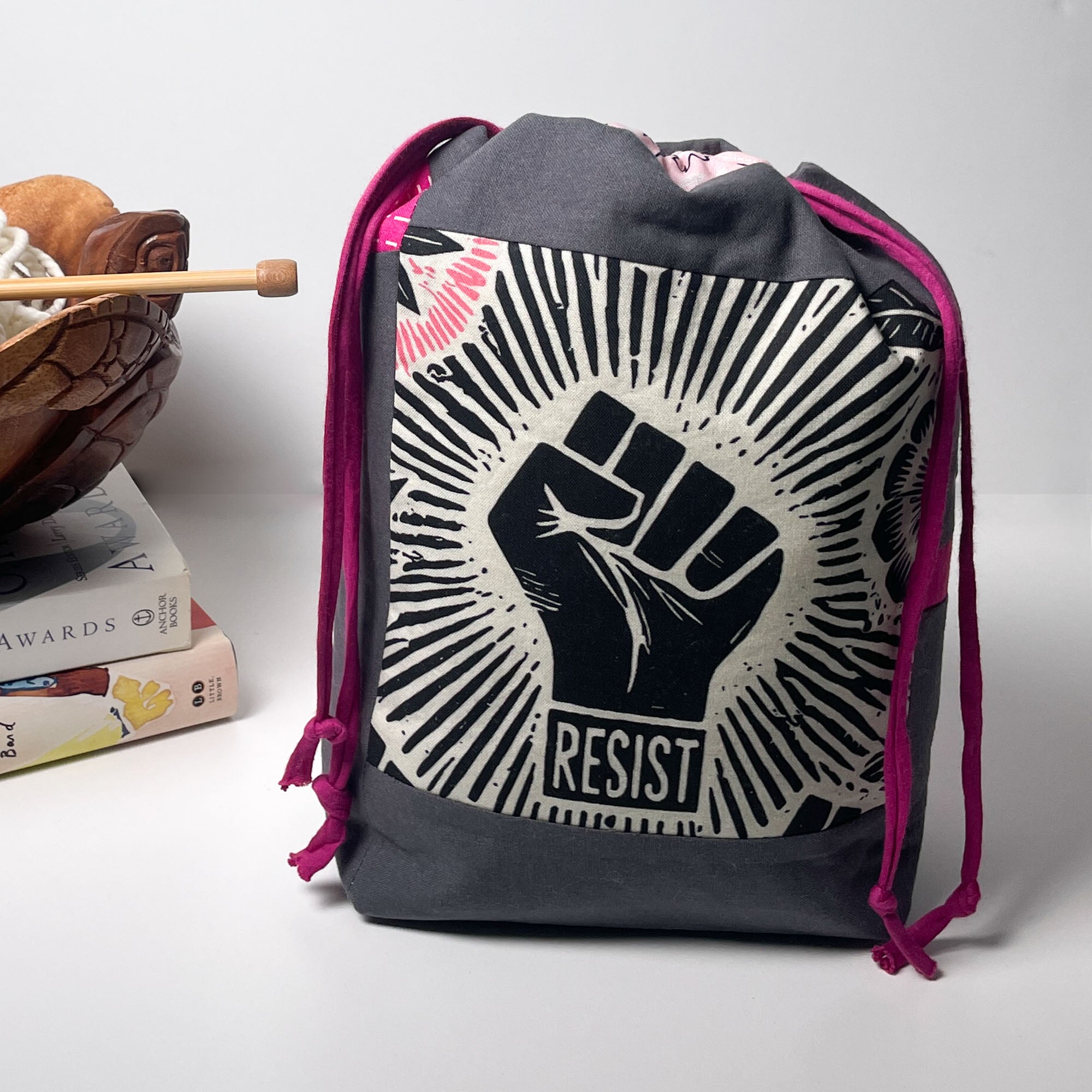 A dark gray drawstring backpack with a prominent black and white graphic of a raised fist and the word 'RESIST' on the front, accented with magenta drawstring cords, set against a light gray background with a decorative book and wooden bowl on the left.