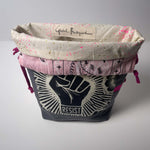 A fabric storage basket or organizer with a dark gray lower section featuring a black and white raised fist graphic and the word 'RESIST,' a pink middle band with black line art, and a cream-colored top lining with pink and gold speckles and a visible 'Global Backyard' label.