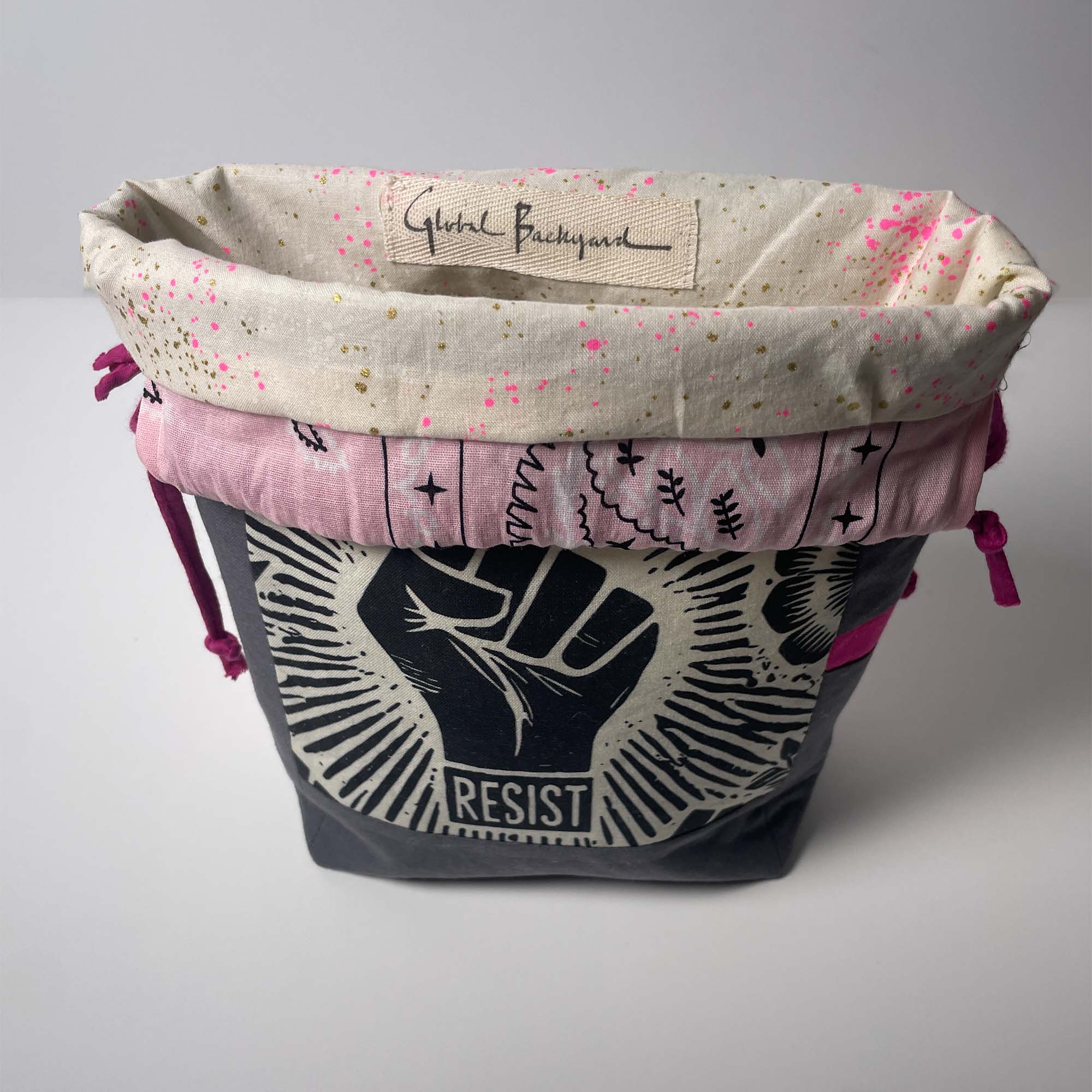 A fabric storage basket or organizer with a dark gray lower section featuring a black and white raised fist graphic and the word 'RESIST,' a pink middle band with black line art, and a cream-colored top lining with pink and gold speckles and a visible 'Global Backyard' label.