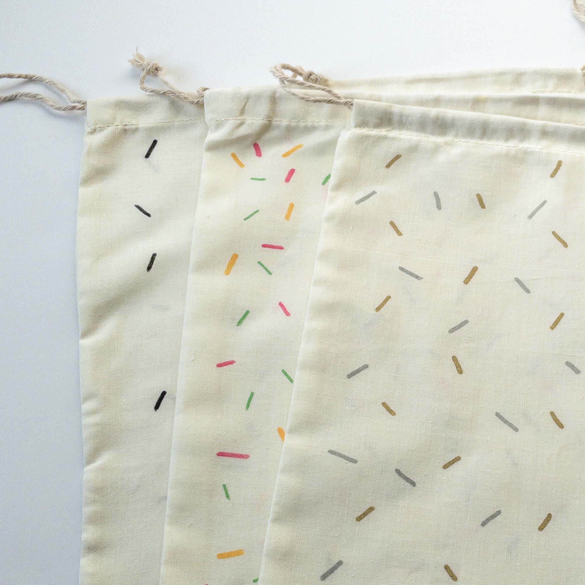 Eco-friendly fabric reusable drawstring pouches in three hand painted confetti patterns -- one in pink, green, and yellow; another in black and white; and the third in metallic gold and silver